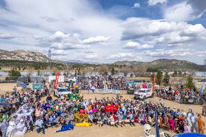Dozens of costumed coffin-race teams gather with their coffins for a group photo, mountains behind.