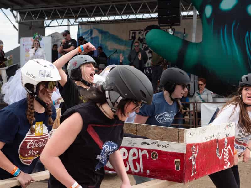 Helmeted racers carry a coffin painted like a beer case at full speed.