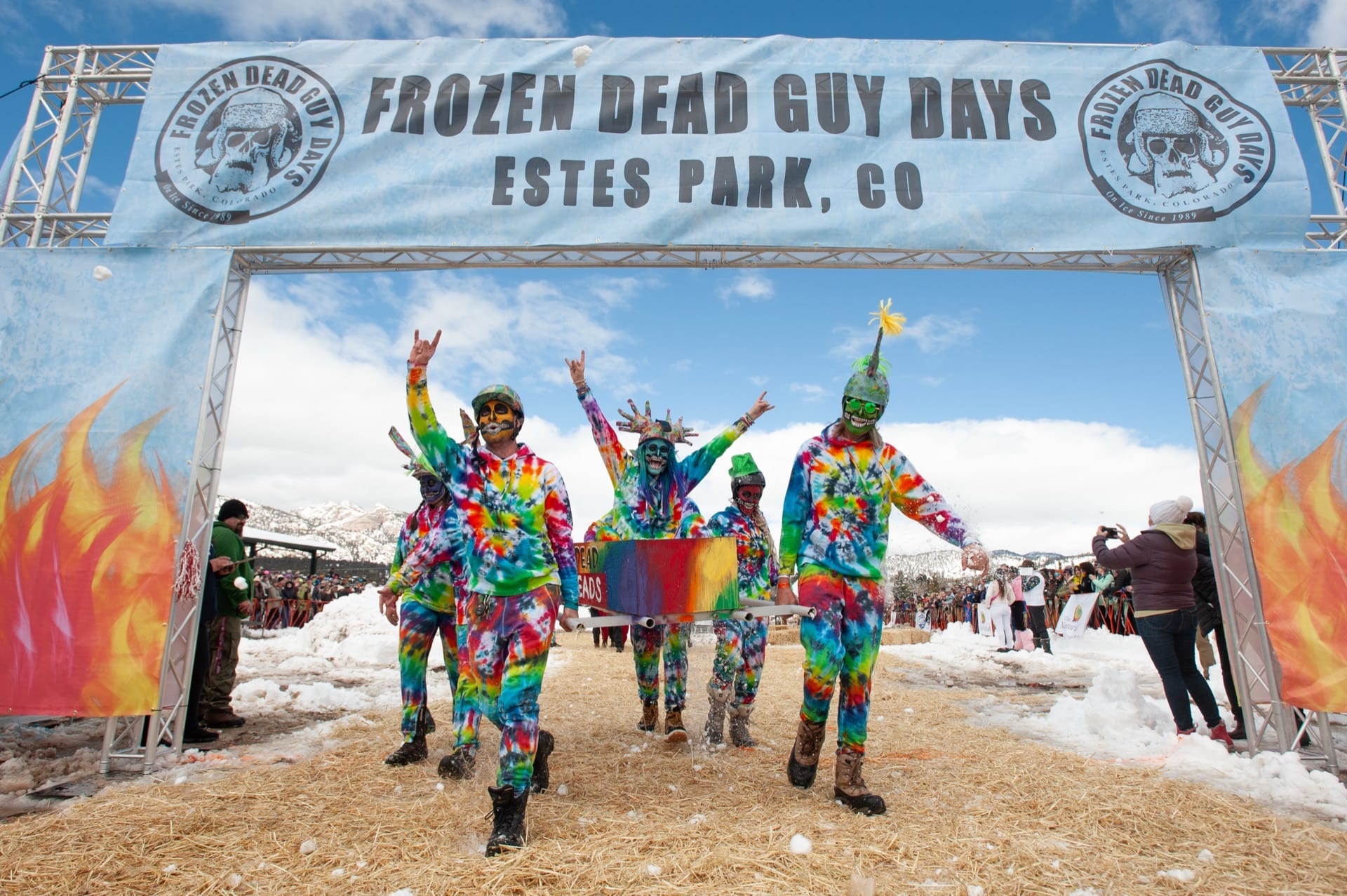 A tie-dye-costumed team walks under the Frozen Dead Guy Days arch on a snowy day.