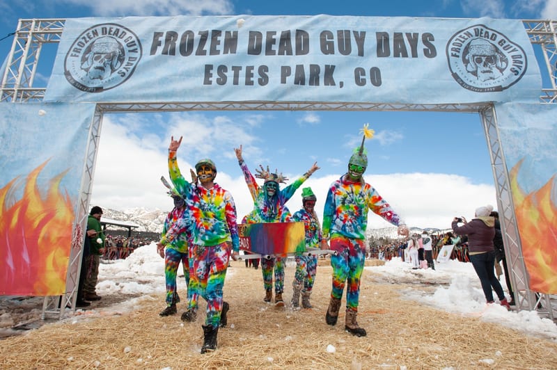 A tie-dye-costumed team walks under the Frozen Dead Guy Days arch on a snowy day.