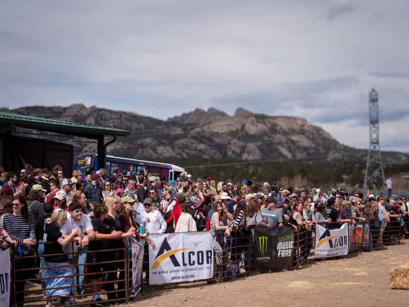 A crowd presses against the course fence beside sponsor banners, peaks in the distance.