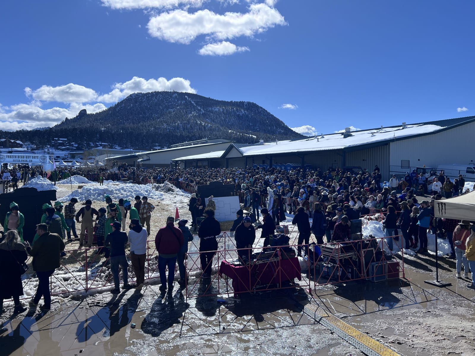Festival-goers gather in the snow with a mountain peak and blue sky behind.