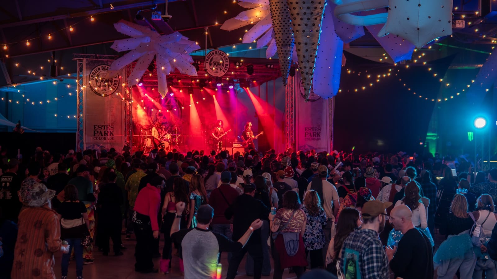 A packed indoor concert under red stage lights and hanging snowflake decorations.