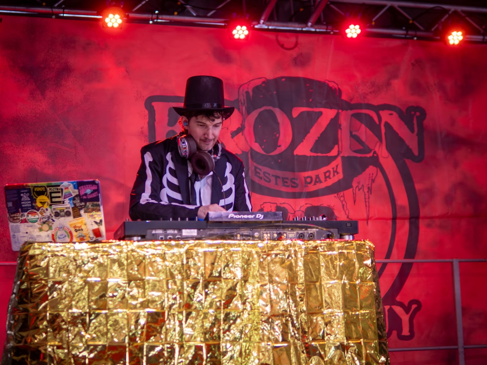 A DJ in a top hat and skeleton suit plays in front of a red Frozen Dead Guy Days backdrop.
