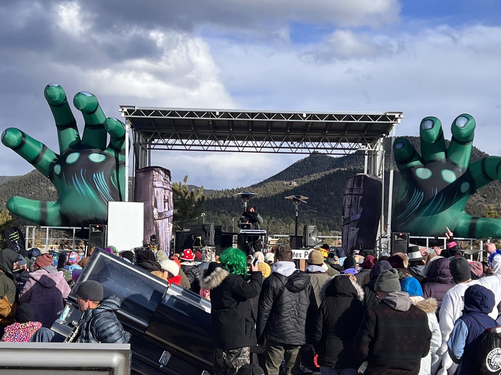 A crowd in winter coats watches the outdoor stage framed by giant zombie hands.