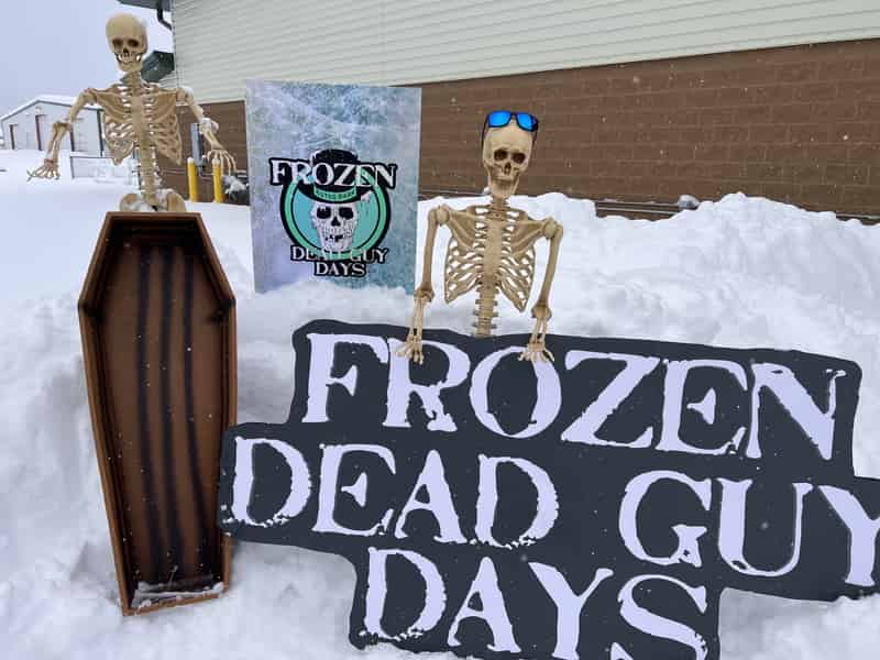 Two skeleton props and a coffin beside a "Frozen Dead Guy Days" sign in deep snow.