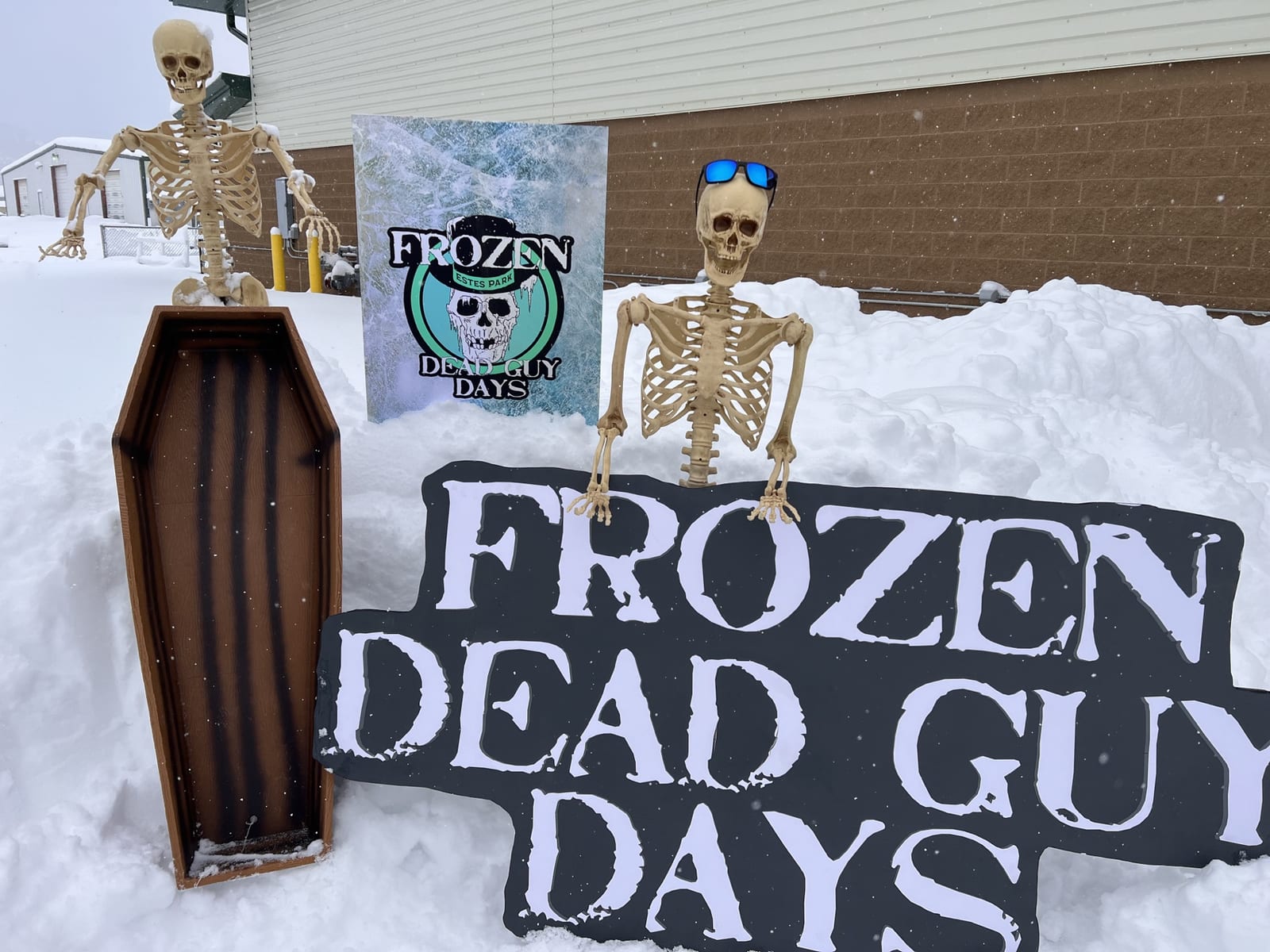 Two skeleton props and a coffin beside a "Frozen Dead Guy Days" sign in deep snow.