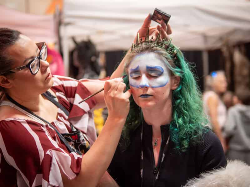 A face painter brushes icy blue skull makeup onto a green-haired woman wearing a spiked crown.