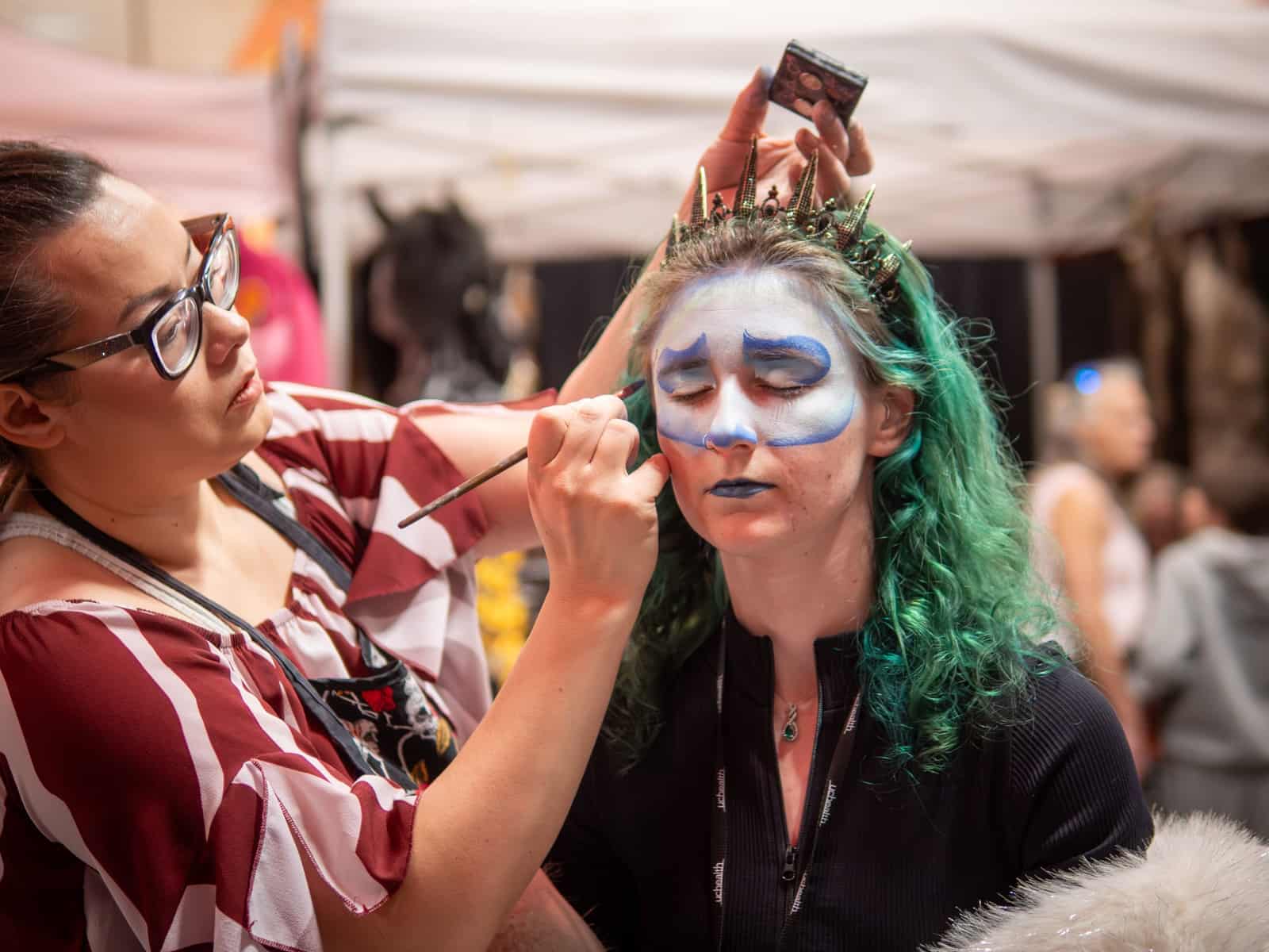A face painter brushes icy blue skull makeup onto a green-haired woman wearing a spiked crown.