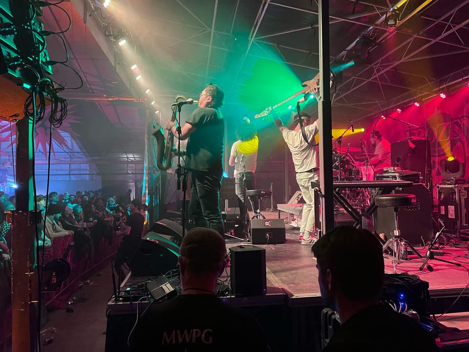 Andy Frasco & The U.N. under green and red light: a sax solo while the guitarist leaps with his guitar raised.