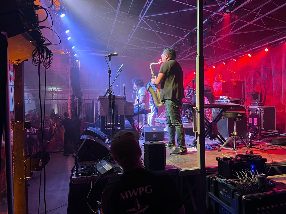 A sax player and the curly-haired keyboardist of Andy Frasco & The U.N. under red and blue light, seen from the side of the stage.