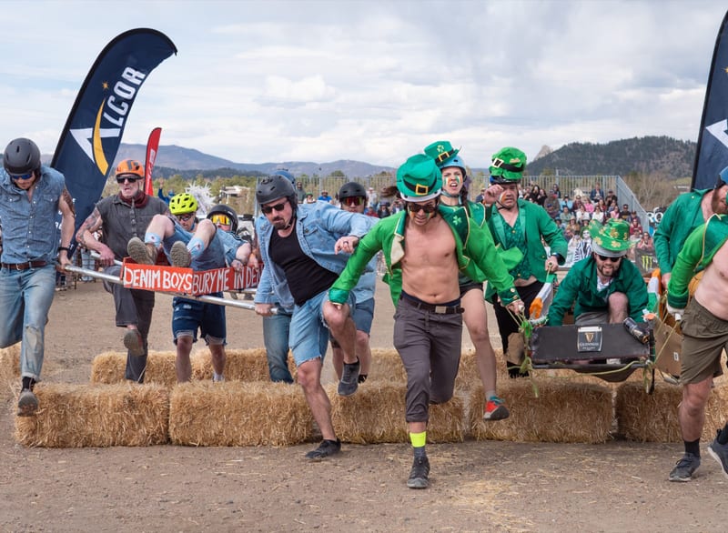 Leprechaun and denim teams leap a row of hay bales side by side, an Alcor flag behind.