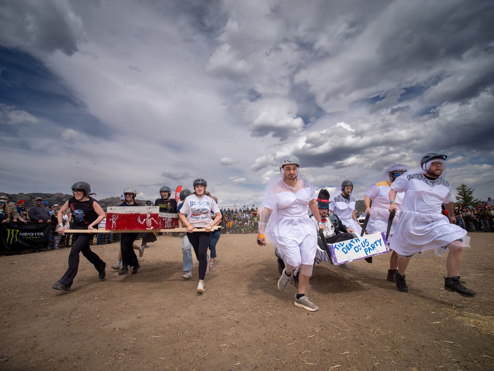 Racers in white wedding dresses and helmets carry a coffin across the dirt course beneath dark clouds.
