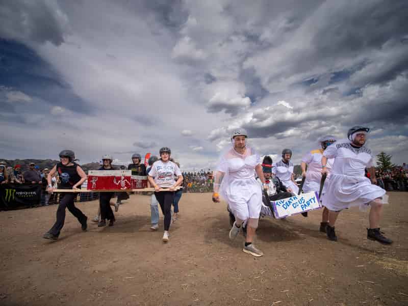 Racers in white wedding dresses and helmets carry a coffin across the dirt course beneath dark clouds.