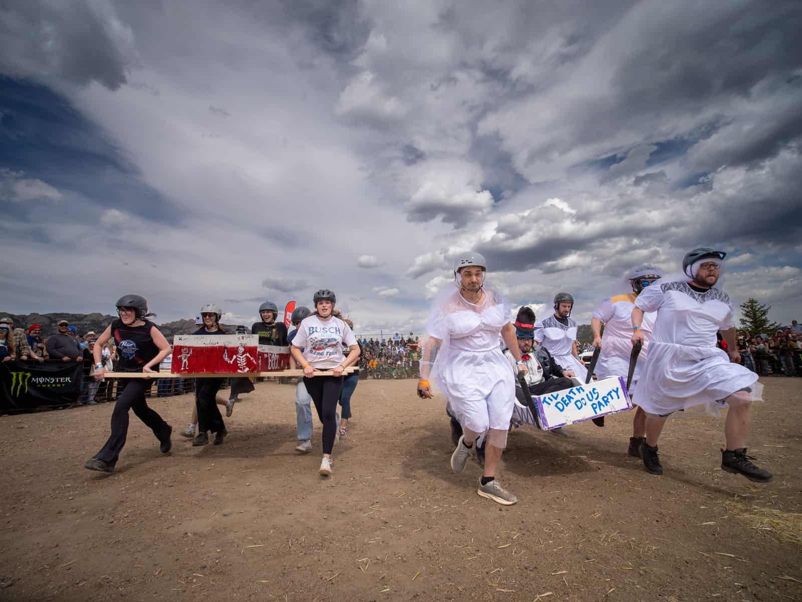 Racers in white wedding dresses and helmets carry a coffin across the dirt course beneath dark clouds.