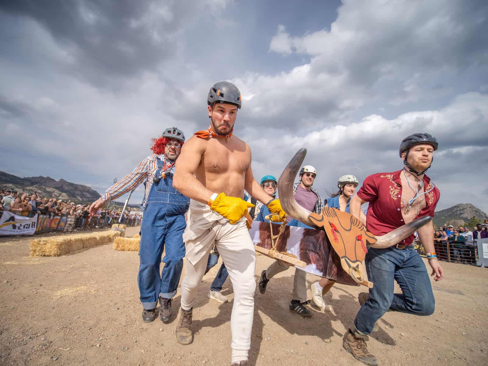 A coffin team carrying a bull-horned coffin charges straight at the camera, faces straining, clouds overhead.
