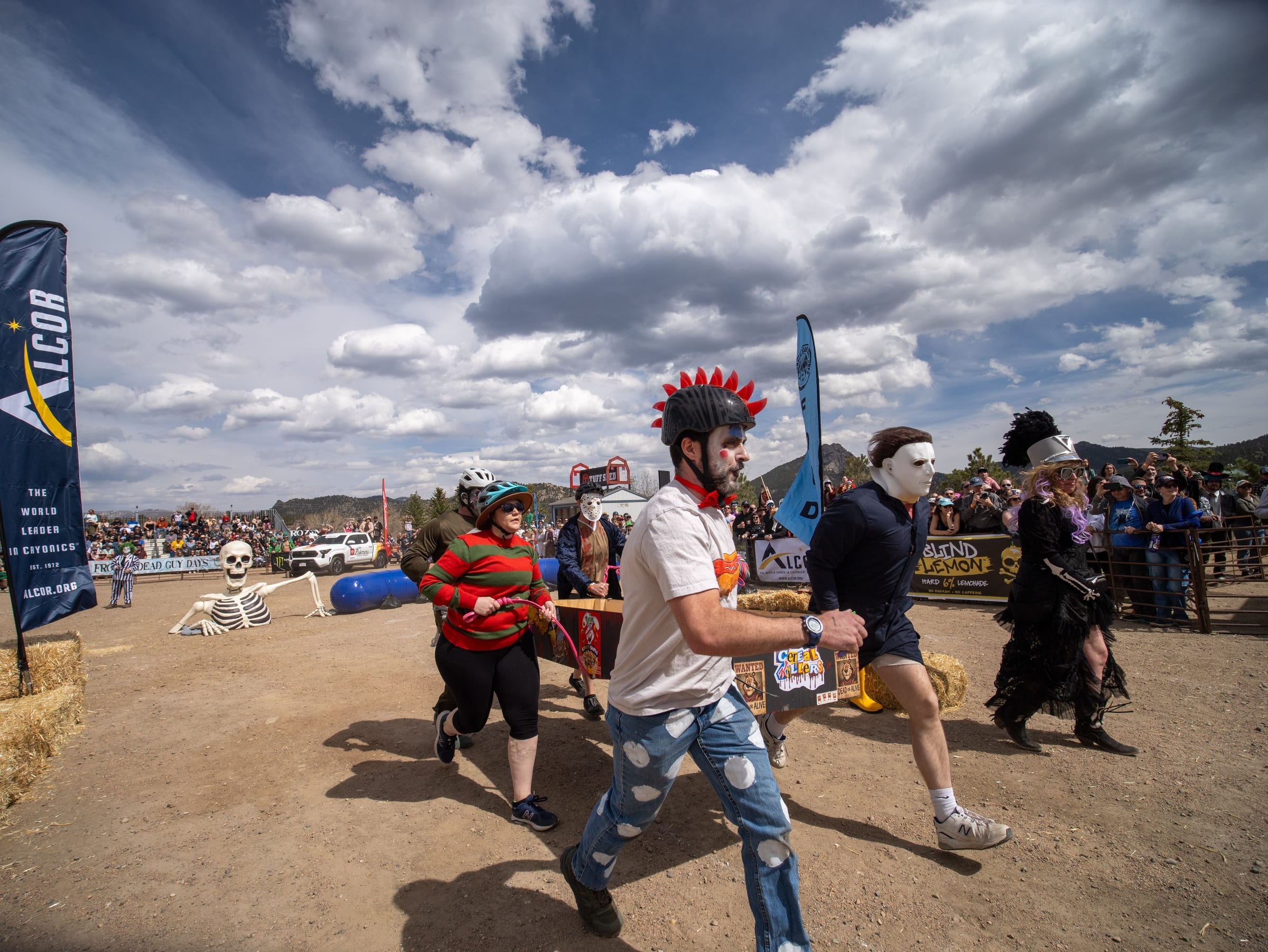 Coffin racers in horror-movie costumes, one in a red mohawk helmet, charge across the course under a huge sky.