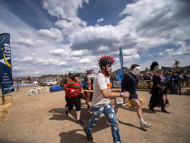 Coffin racers in horror-movie costumes, one in a red mohawk helmet, charge across the course under a huge sky.