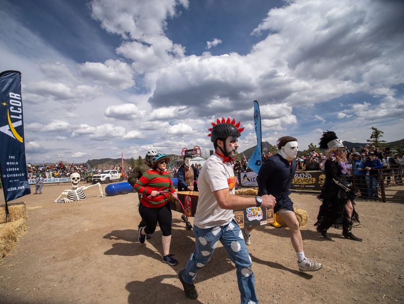 Coffin racers in horror-movie costumes, one in a red mohawk helmet, charge across the course under a huge sky.
