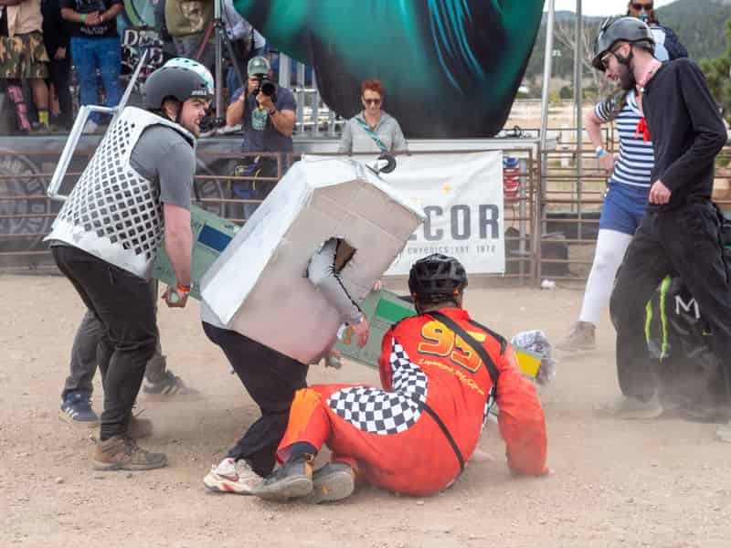 A coffin comes apart mid-race as a racer in a checkered suit lands on the dirt.