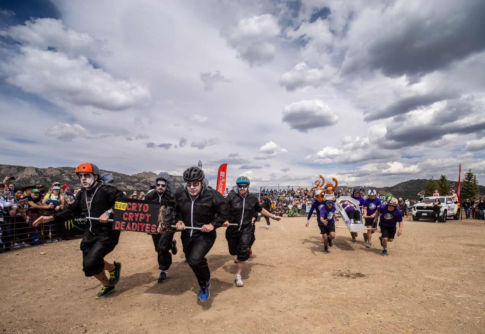 A team in black jumpsuits and pale corpse makeup races a "Cryo Crypt Deadites" coffin under cloudy skies.