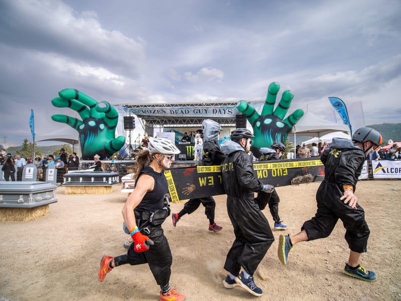 A team in black racing suits and helmets runs its coffin past the Frozen Dead Guy Days stage and its giant green inflatable hands.