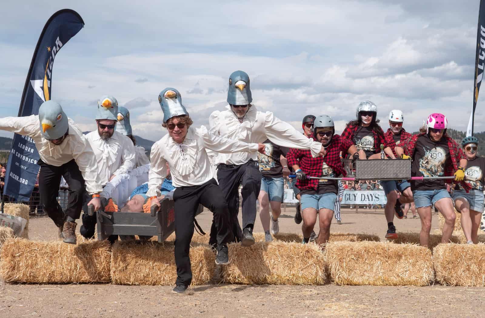 Two coffin teams, one in pigeon-head helmets, leap a row of hay bales mid-race.