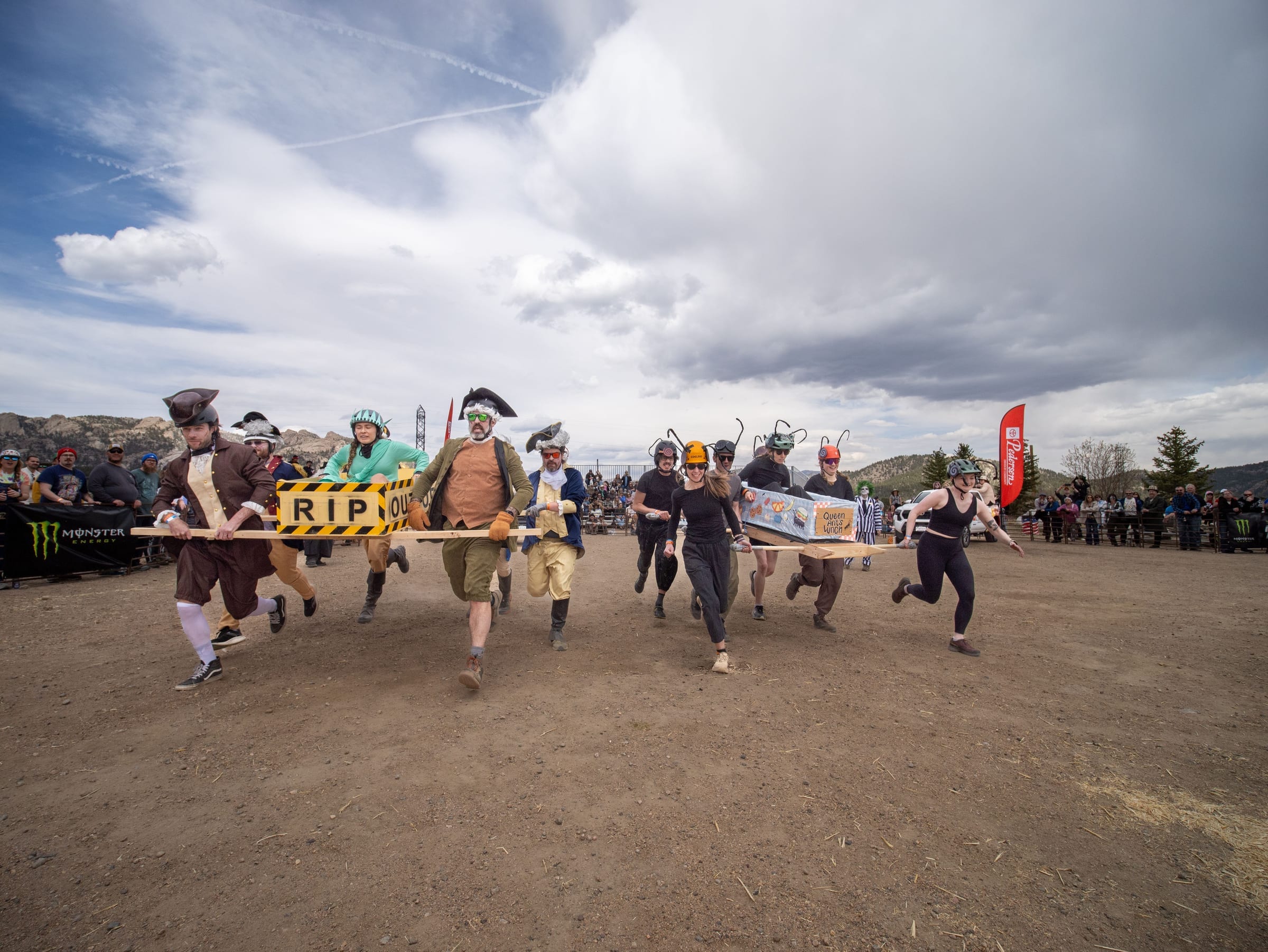 Two coffin teams sprint off the start side by side, one in colonial costumes with an "RIP" coffin, under dramatic clouds.