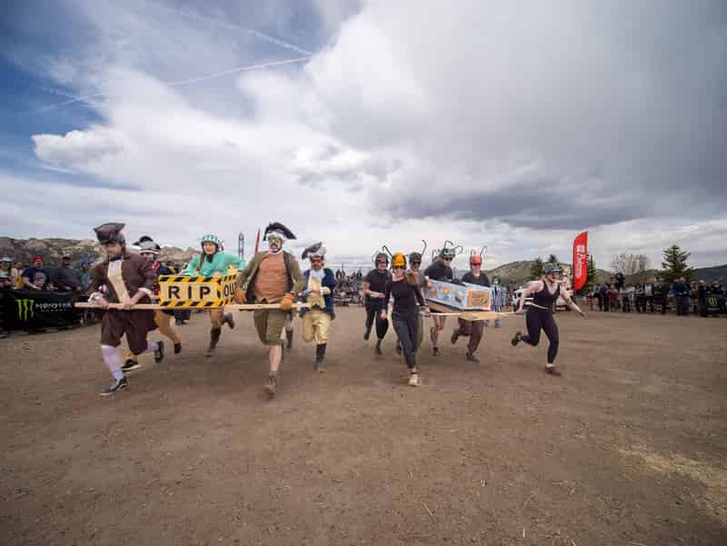 Two coffin teams sprint off the start side by side, one in colonial costumes with an "RIP" coffin, under dramatic clouds.