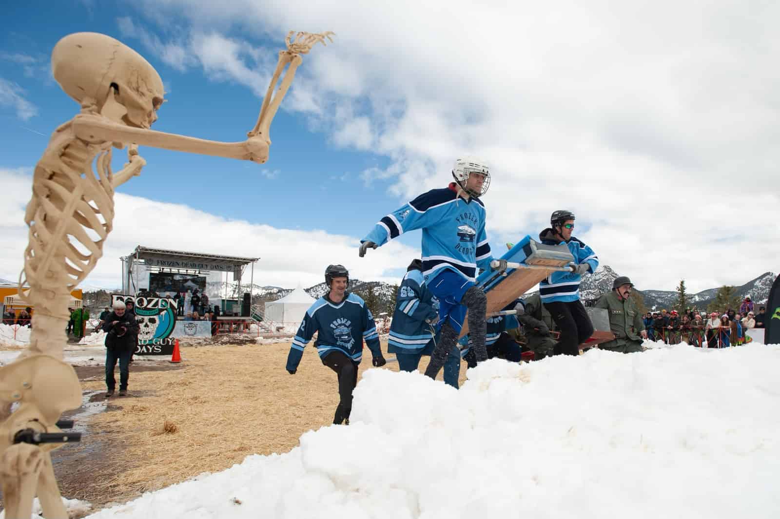 A team in blue hockey jerseys carries a coffin over a snowbank as a skeleton prop waves in front.