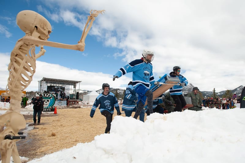 A team in blue hockey jerseys carries a coffin over a snowbank as a skeleton prop waves in front.