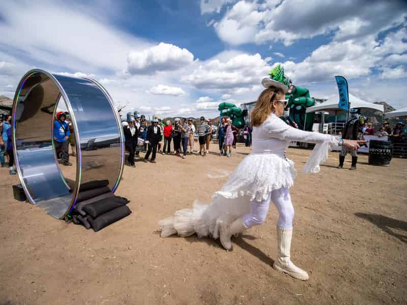 A performer in a white feathered gown gestures across the race course beside a giant mirrored wheel.