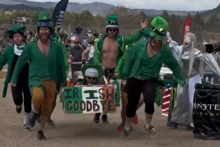 The Irish Goodbye team, in green leprechaun coats and hats, charges toward the camera carrying their IRISH GOODBYE coffin.