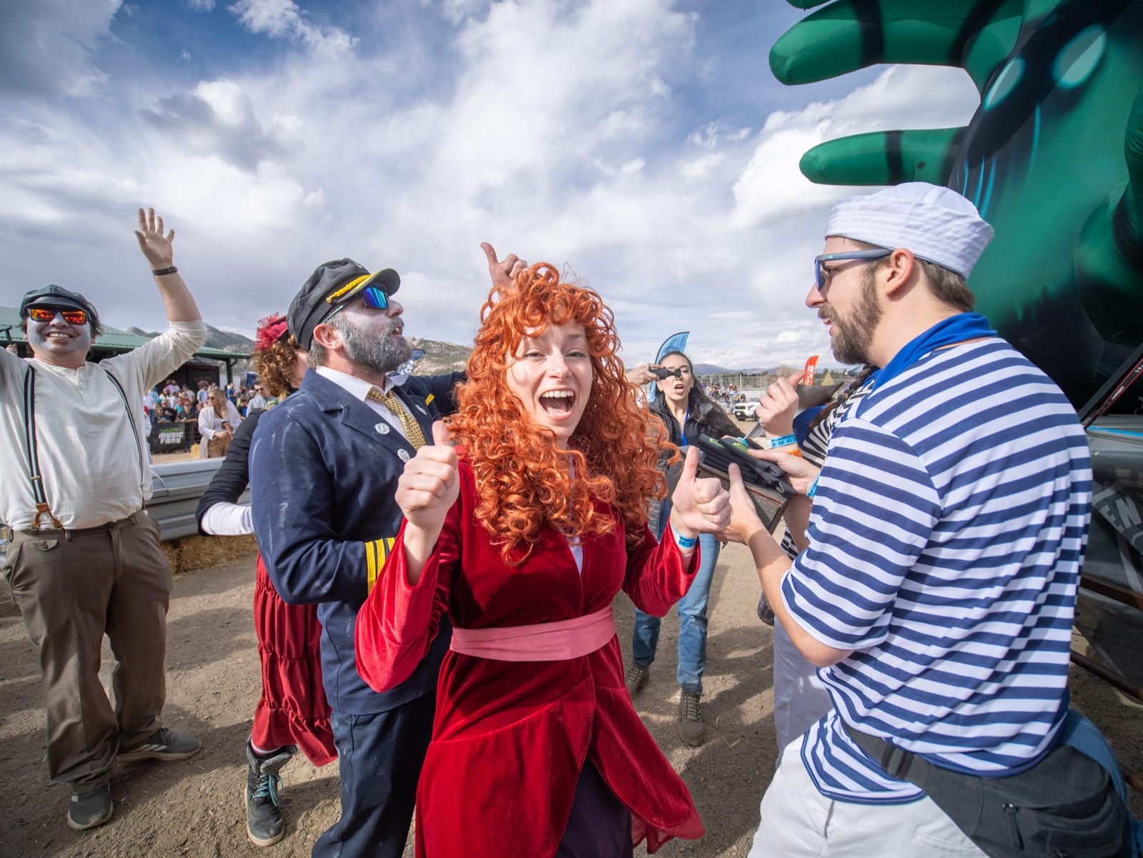 A red-haired racer in a red gown laughs with a teammate in a striped sailor shirt beside the course.