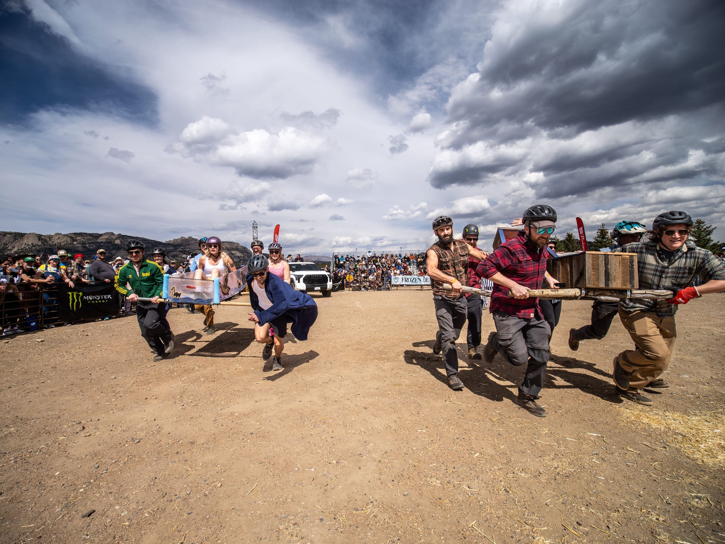Two costumed coffin teams sprint toward the camera under a vast stormy sky, a crowd lining the course.