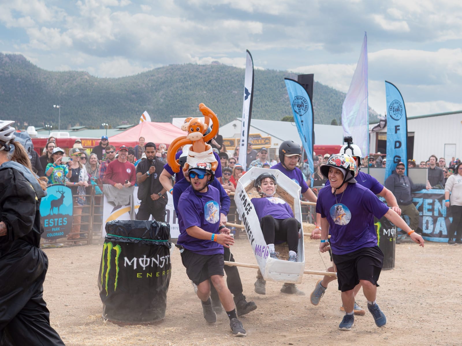 A team in purple shirts races a coffin past a Monster Energy barrel.