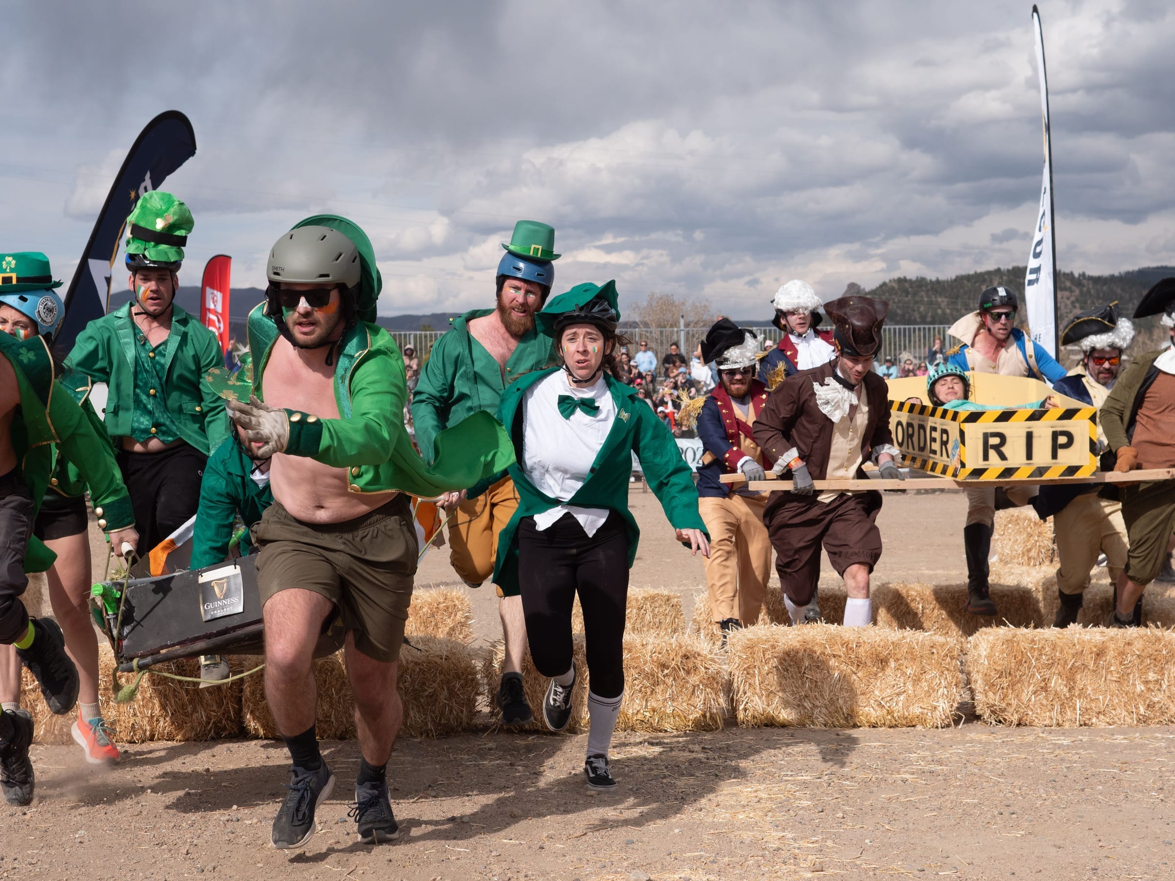 A leprechaun team and a colonial-costumed team carry their coffins over a row of hay bales, side by side.