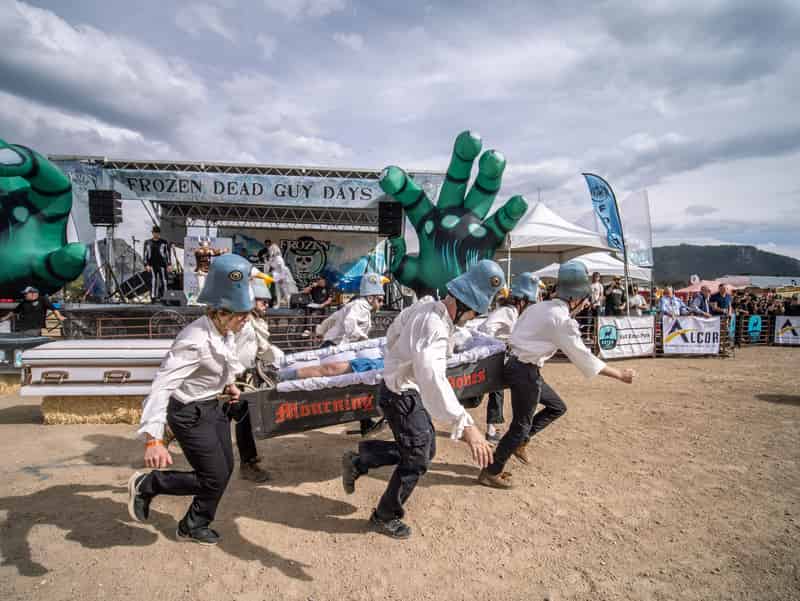 Pigeon-helmeted racers rush an open coffin past the Frozen Dead Guy Days stage and giant green hands.