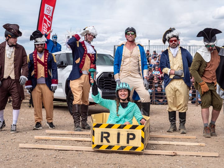 A team in colonial costumes grins beside a teammate riding in an RIP coffin, a Pedersen Toyota flag and truck behind.