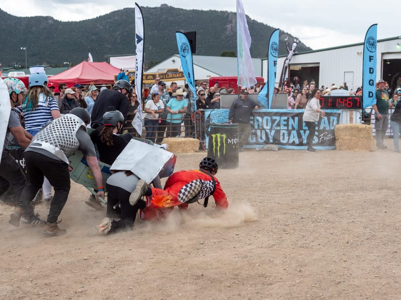 Racers pile up in the dust as a coffin tips over mid-race, a racer in a checkered suit sprawled on the ground.