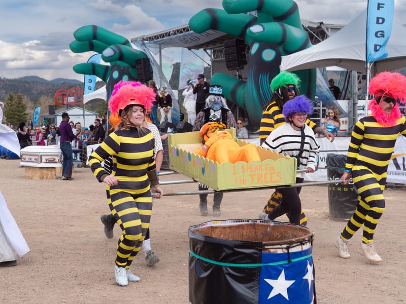 A team in yellow-and-black bee stripes and rainbow wigs races a coffin past a star-painted barrel.