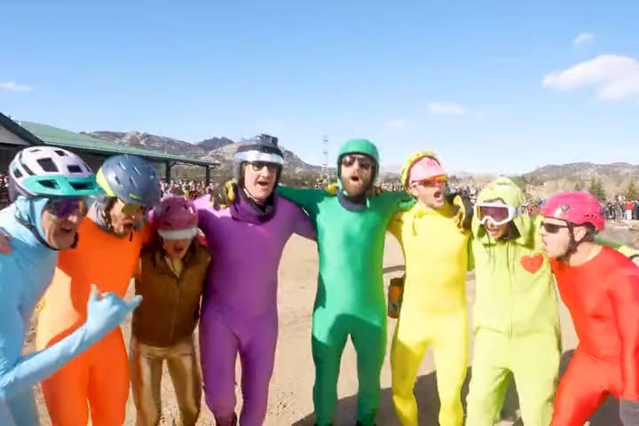 The Rainbros, in orange, gold, purple, green, yellow, lime and red bodysuits and helmets, stand arm in arm at the 2023 Coffin Races.