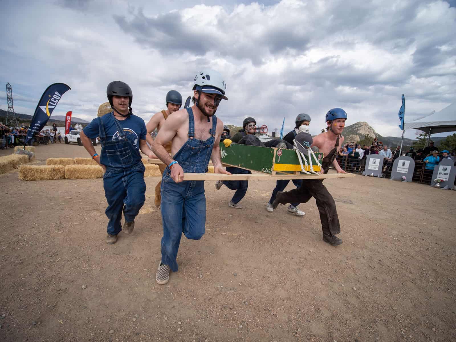 Racers in denim overalls carry a green coffin with a skeleton passenger past hay bales and mountain peaks.