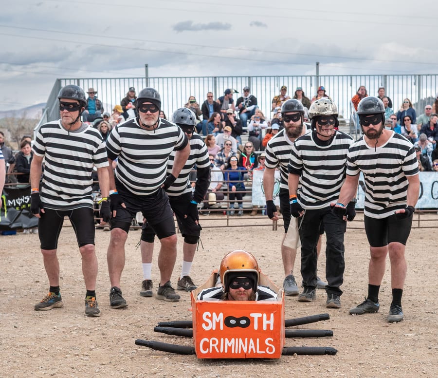 The Smooth Criminals, six pallbearers in black-and-white convict stripes, stand behind their orange coffin with their helmeted corpse lying inside.
