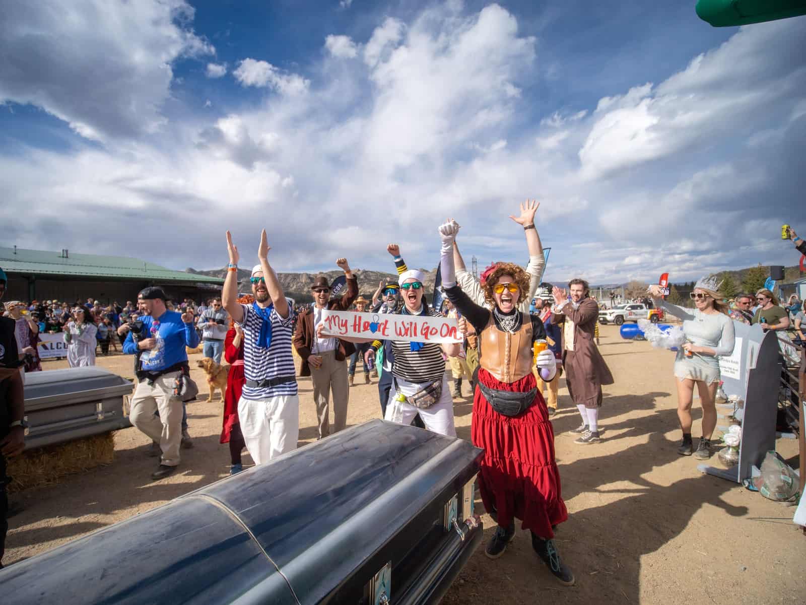 Costumed racers throw their arms up and cheer behind a black casket under a bright, cloud-streaked sky.