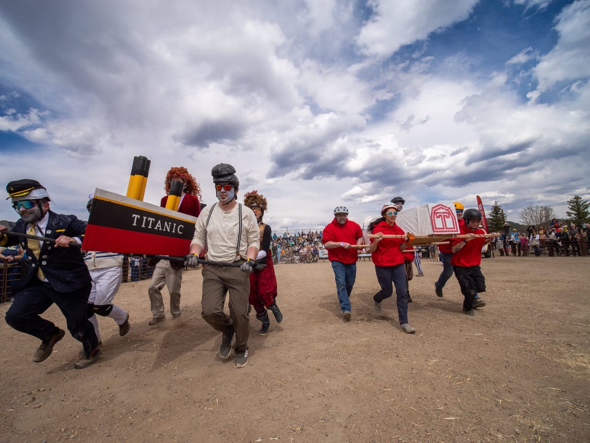 Two coffin teams race side by side under a huge Colorado sky: a ship-captain team carrying a Titanic coffin and a team in red shirts.