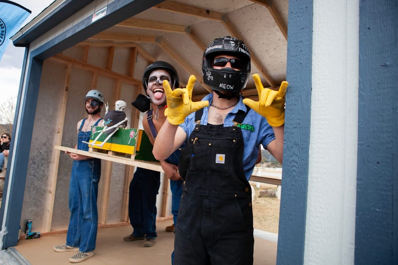 Racers in overalls and helmets wait inside the Tuff Shed at the start, one throwing horns in yellow work gloves.