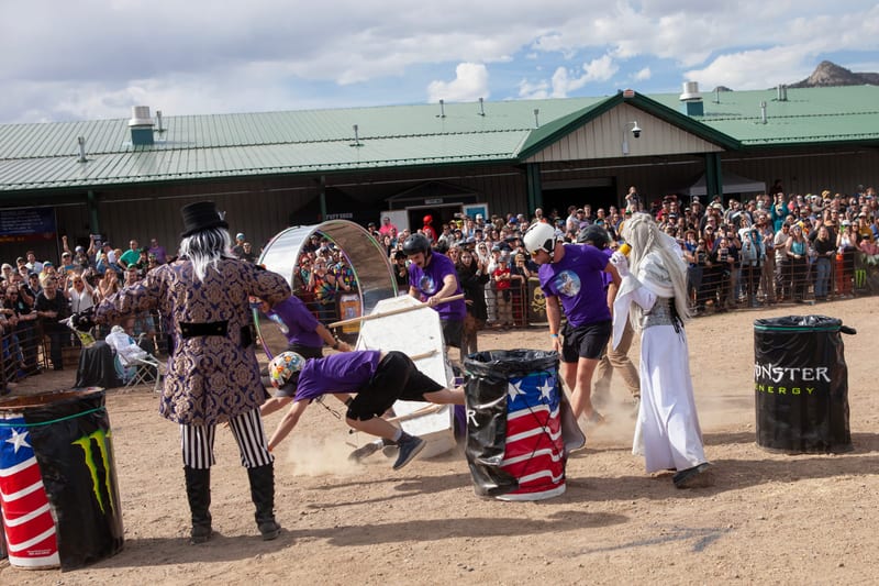 A racer in purple tumbles headfirst as a coffin crashes beside the Monster Energy barrels, costumed characters looking on.
