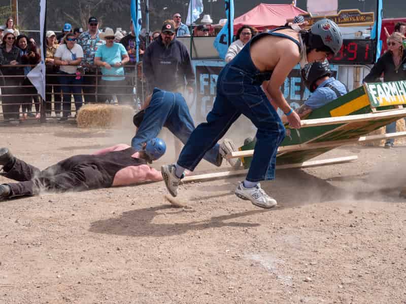 A racer in overalls hurdles a teammate who has gone face-first into the dirt, their coffin tipping beside them.