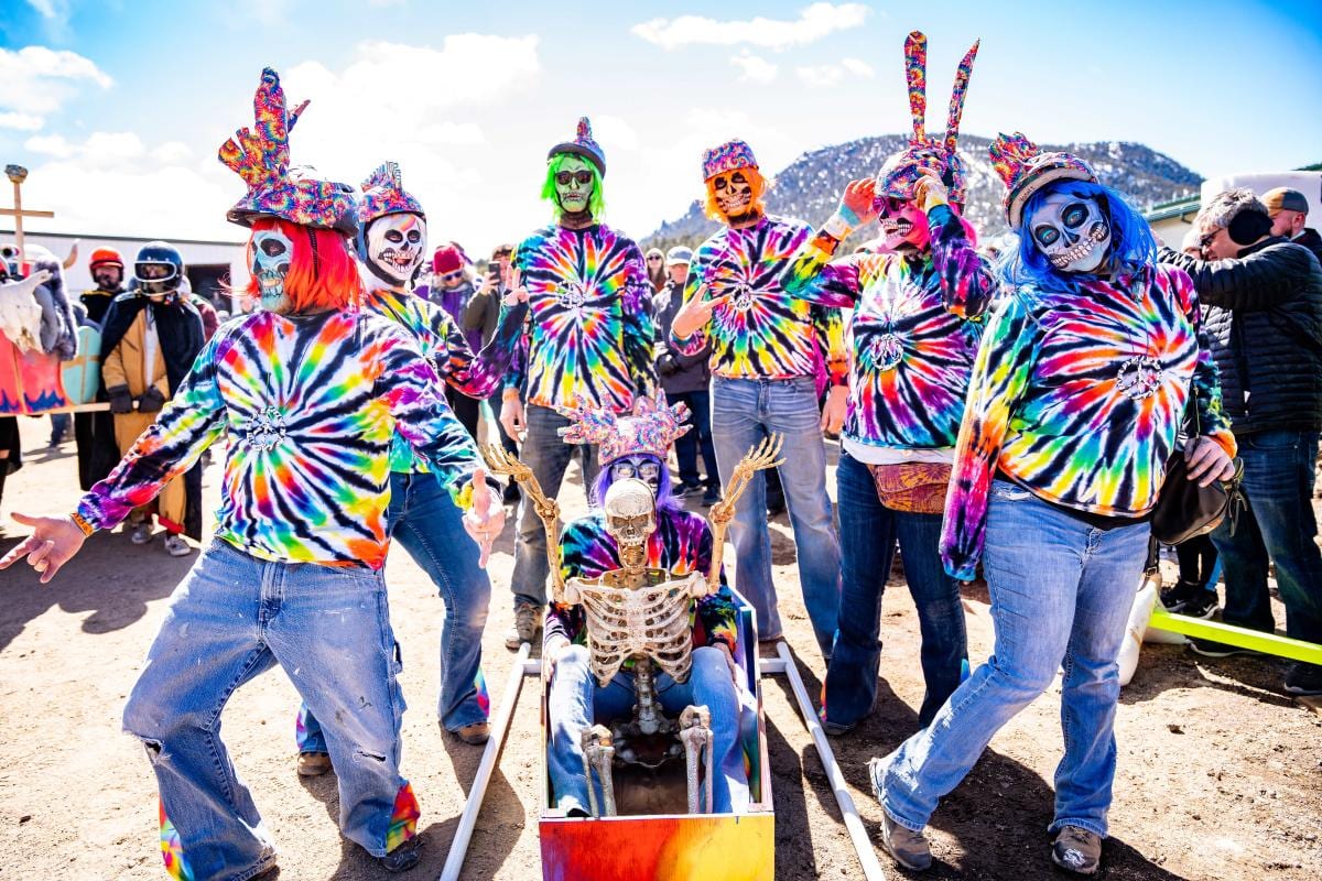 A coffin team in tie-dye shirts and painted skull masks poses around a coffin carrying a skeleton, a mountain behind.